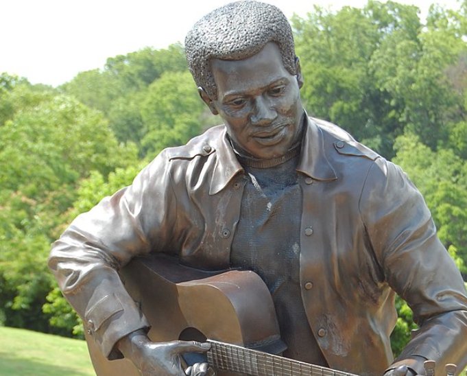 Bronze statue of a man playing guitar, wearing a jacket, against a backdrop of greenery.