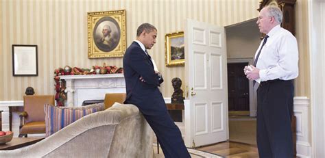 A discussion between two men in the Oval Office, with one leaning against a couch and the other standing with a notepad. The room is decorated for the holidays.