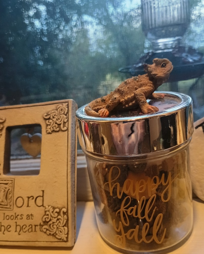 A decorative glass jar labeled 'happy fall y'all' filled with various autumn-themed items, with a small lizard figurine perched on top and a photo frame in the background.